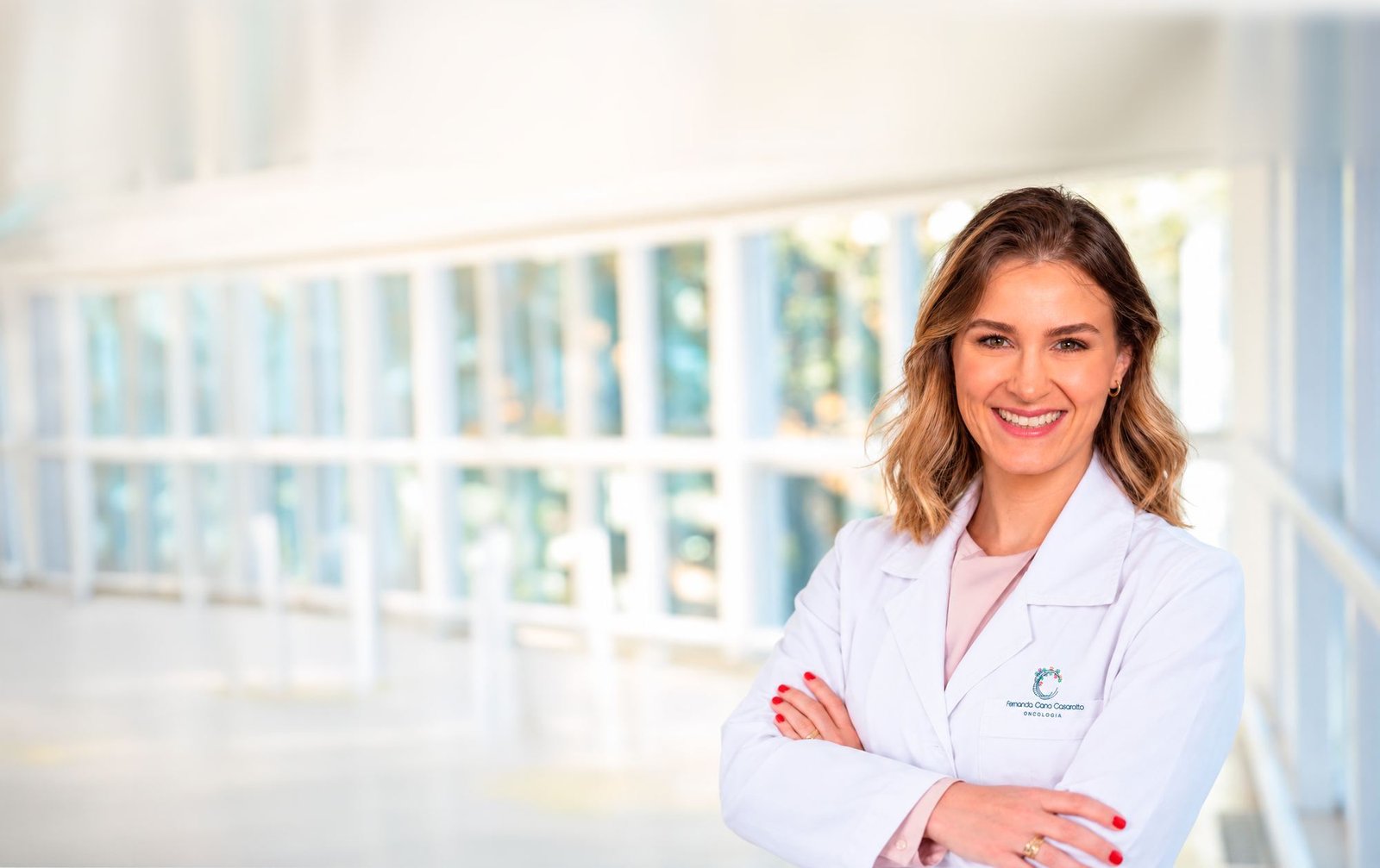 Dra. Fernanda Cano Casarotto, de jaleco branco e braços cruzados, em um corredor iluminado do hospital